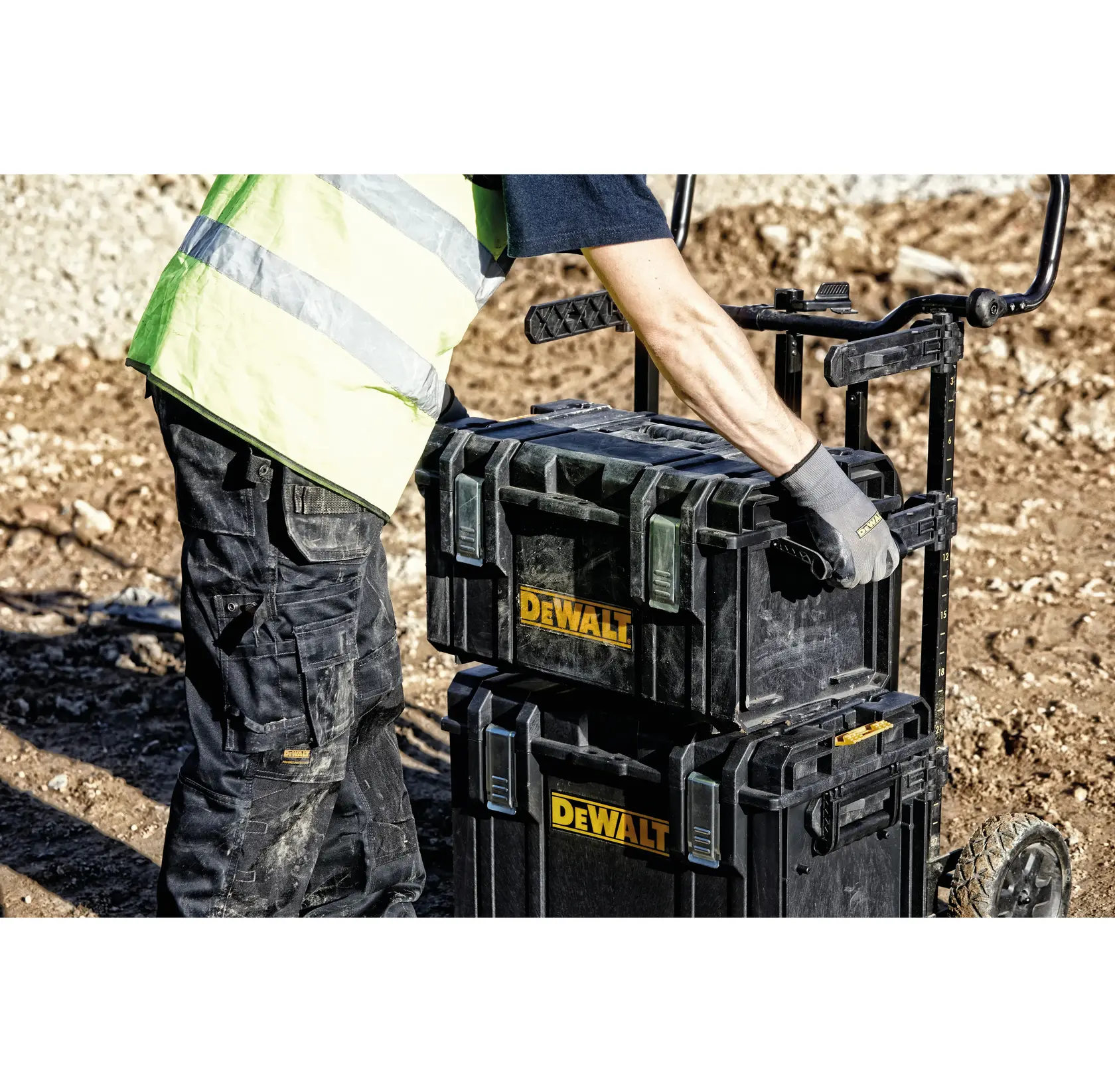 Uma pessoa usando colete de alta visibilidade e luvas de trabalho move caixas de ferramentas DEWALT empilhadas em um carrinho em um canteiro de obras.