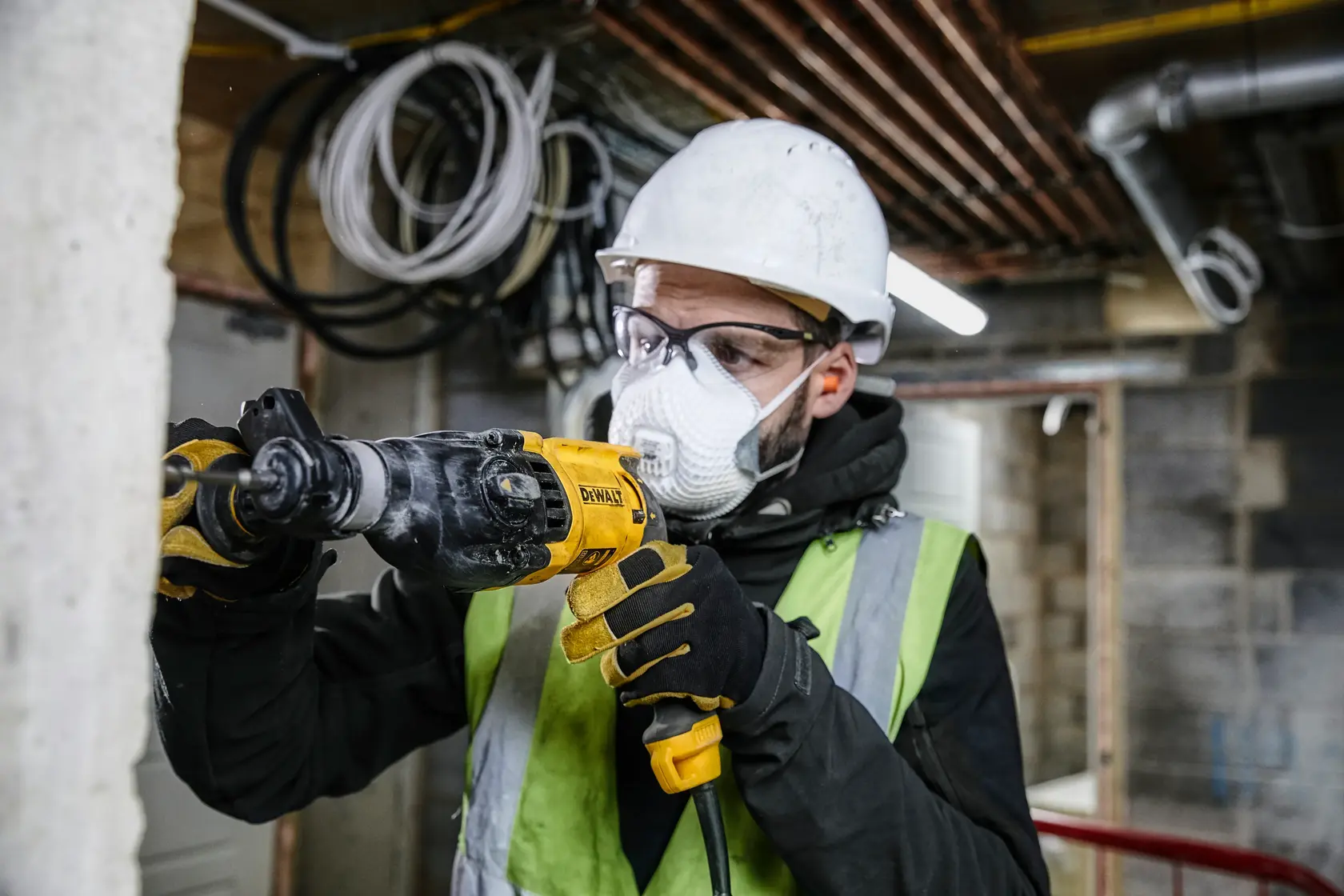 Uma pessoa com capacete de segurança, luvas e colete refletivo está usando uma furadeira DEWALT para trabalhar em uma parede de concreto em um ambiente de construção. O rosto está desfocado por privacidade.
