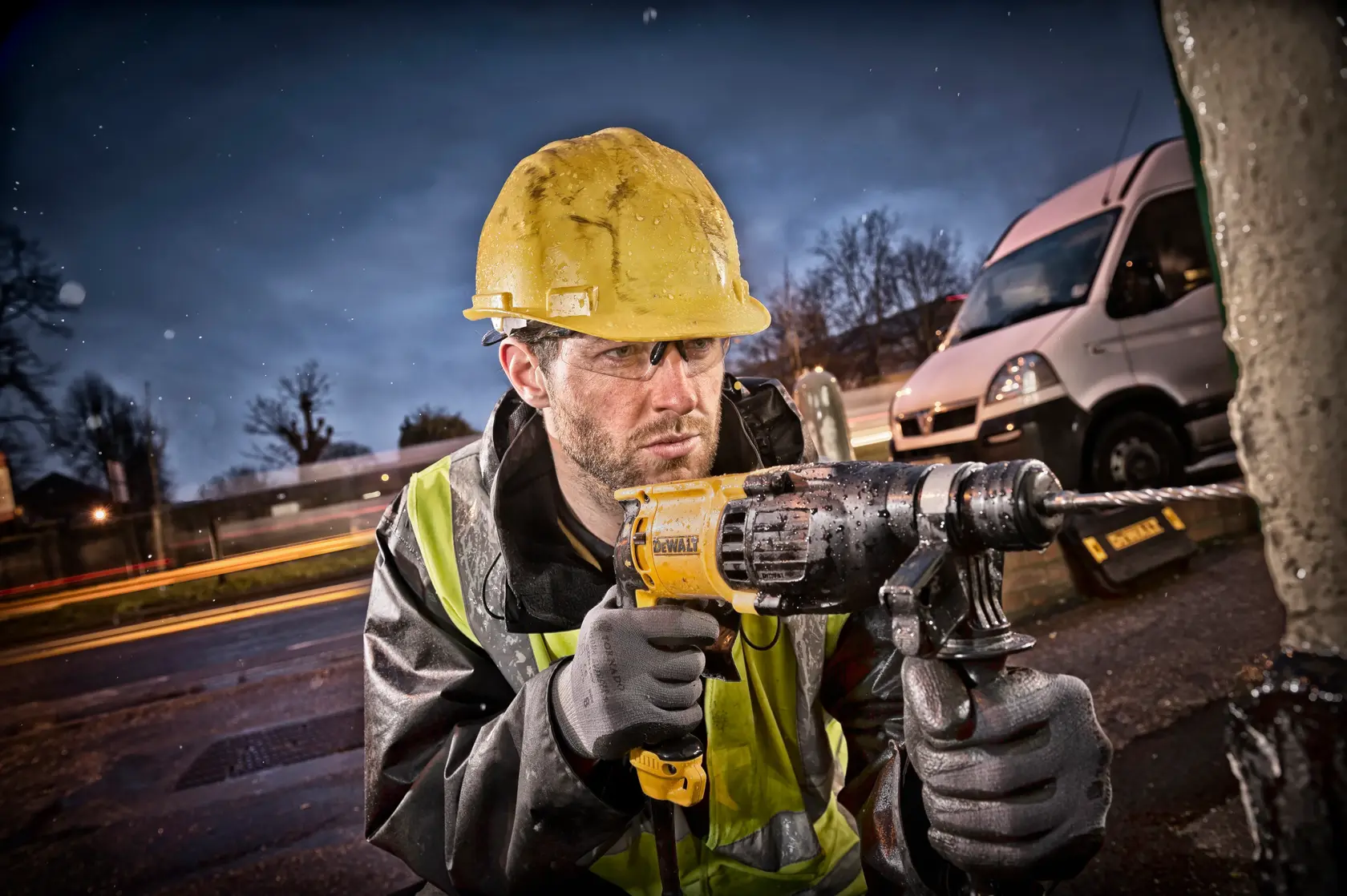 Um trabalhador da construção usando capacete amarelo e colete refletivo utiliza uma furadeira de impacto DEWALT ao ar livre, com uma van branca e luzes desfocadas ao fundo.
