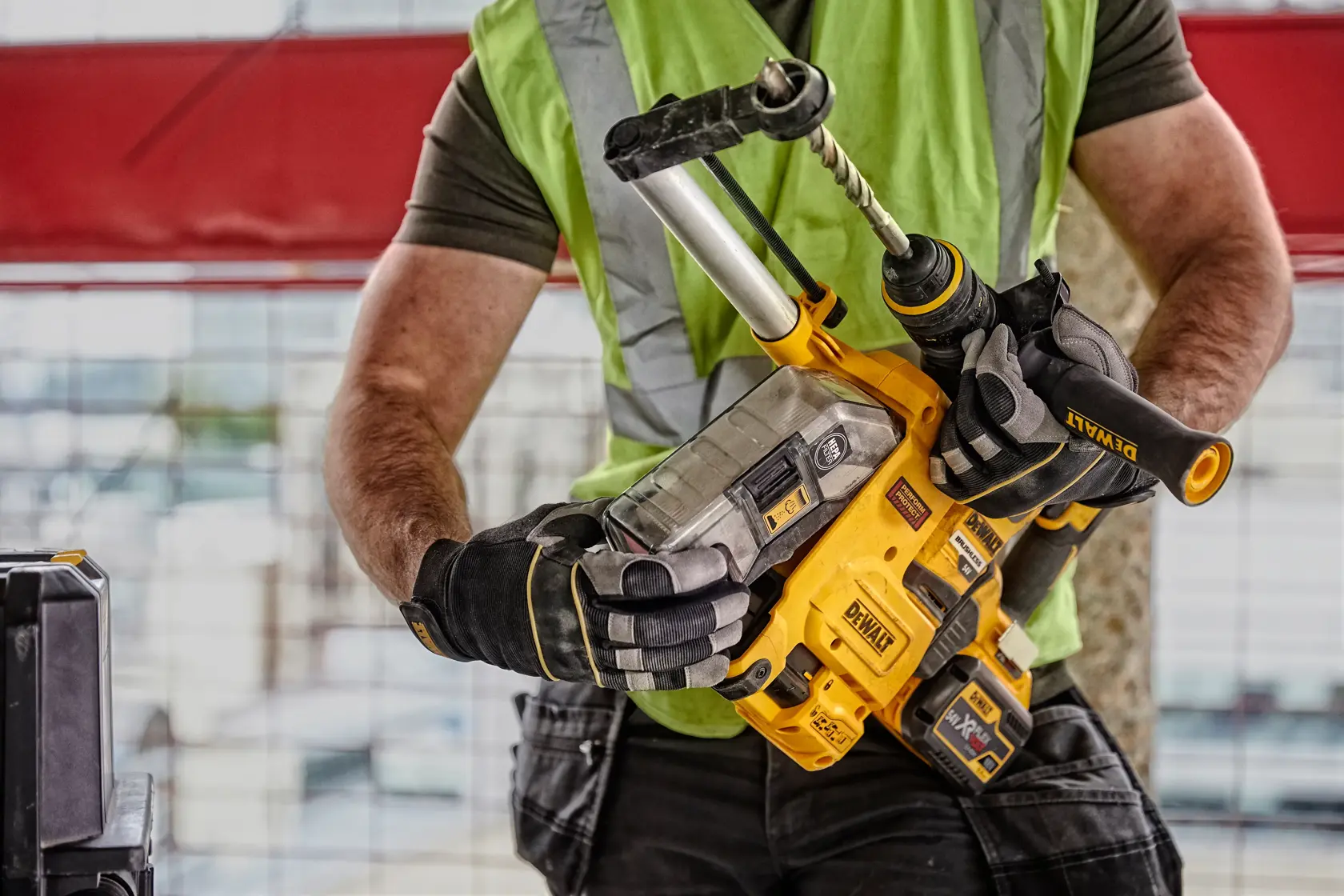 Uma pessoa usando colete de segurança verde e luvas segura uma furadeira de impacto sem fio DEWALT D25304DH com sistema de extração de poeira acoplado em um canteiro de obras.