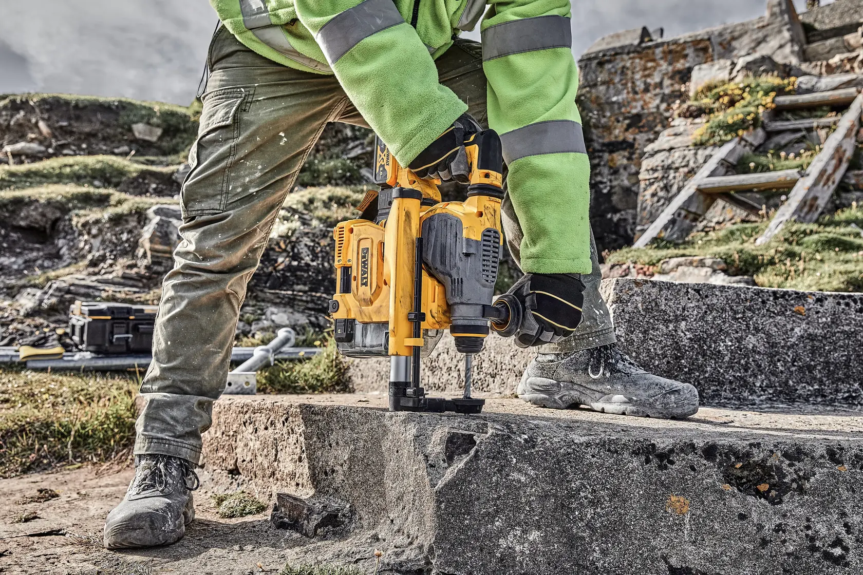 Pessoa usa furadeira DEWALT no concreto ao ar livre, com luvas, botas e equipamento de segurança.