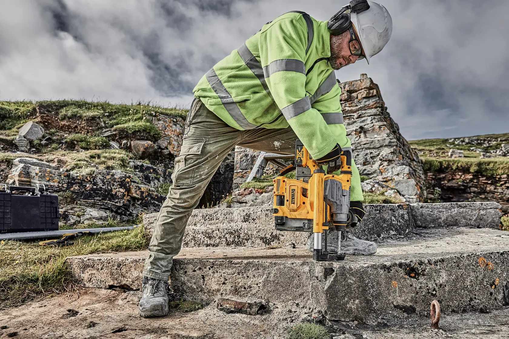 Trabalhador fura concreto ao ar livre com ferramenta DEWALT amarela, usando equipamento de segurança e jaqueta.