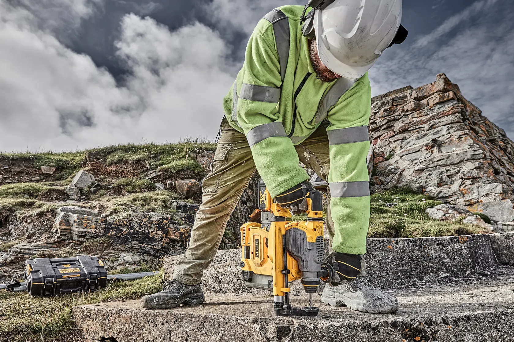 Trabalhador usa ferramenta DEWALT no concreto ao ar livre, com EPI e capacete, caixa de ferramentas ao lado.
