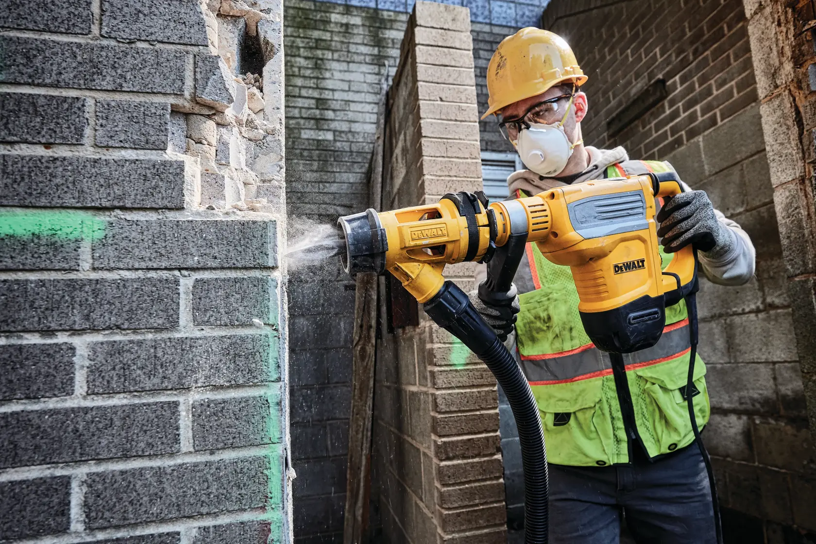 Um trabalhador da construção civil com capacete amarelo e colete de segurança utiliza um martelete rotativo DEWALT (modelo D25614K) para perfurar uma parede de tijolos, com poeira sendo expelida por um sistema de extração de poeira acoplado.