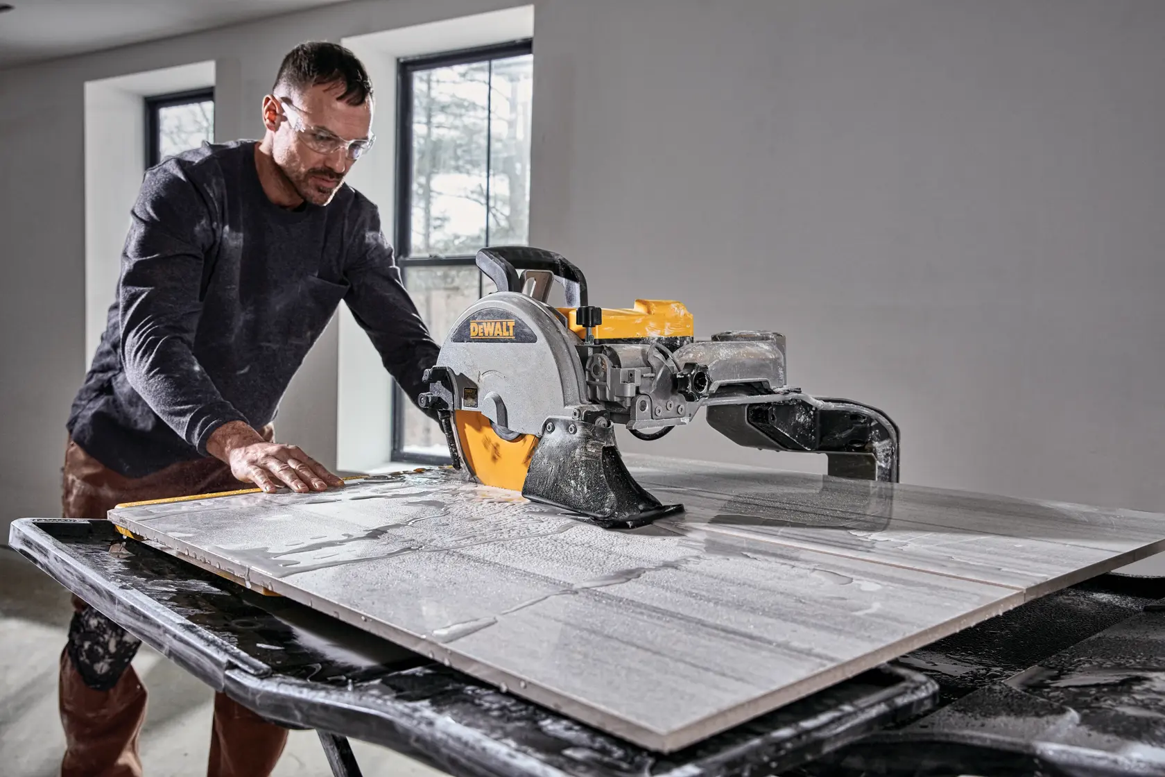 Uma pessoa usa uma serra de azulejos DEWALT D36000 para cortar um grande azulejo em uma mesa de trabalho em uma sala com duas janelas.