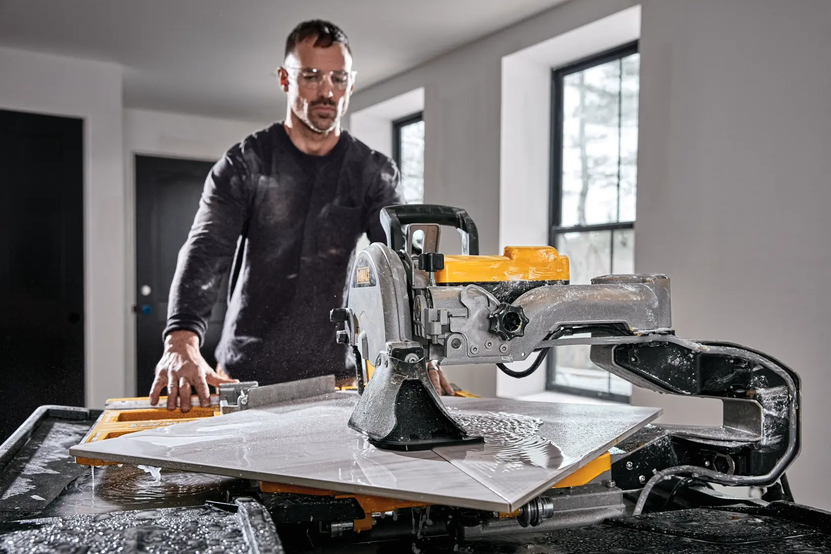 Uma pessoa operando uma serra de azulejo úmida DEWALT D36000-QS em um cômodo bem iluminado, cortando um azulejo grande com água respingando na superfície de trabalho.