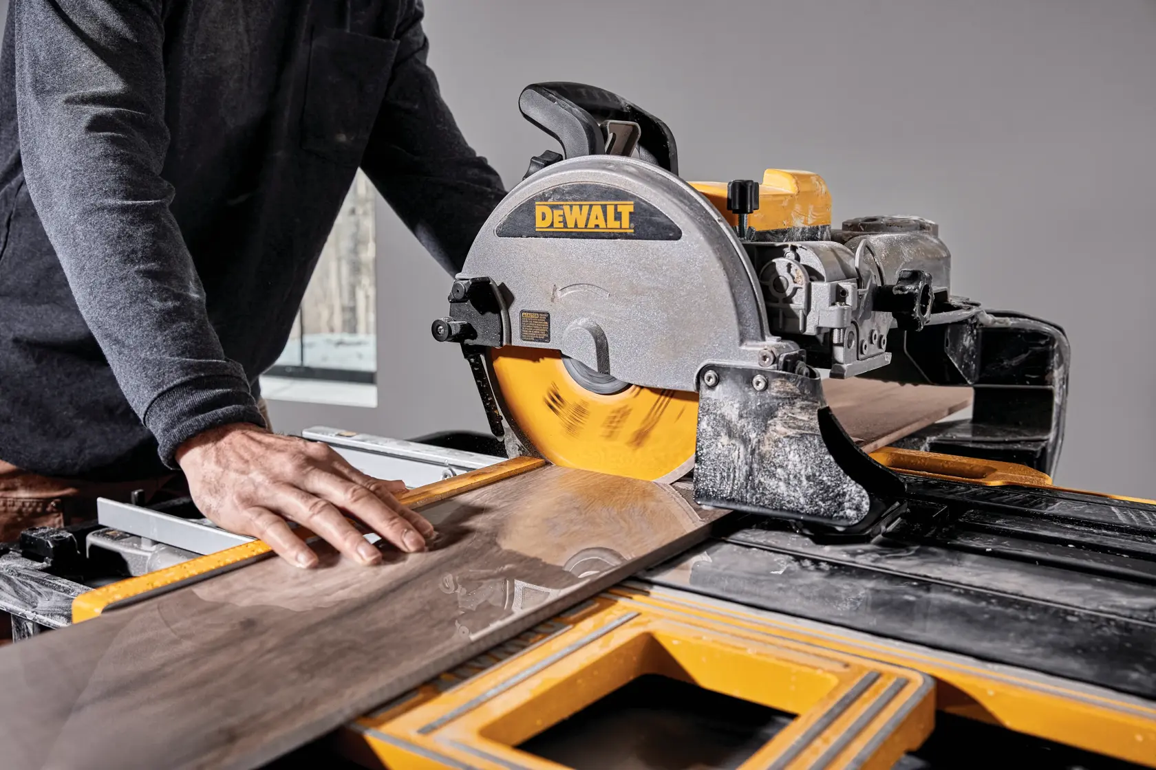Uma pessoa está cortando uma peça grande de azulejo com uma serra para azulejos DEWALT. A lâmina da serra está girando e a pessoa guia o azulejo cuidadosamente com a mão.