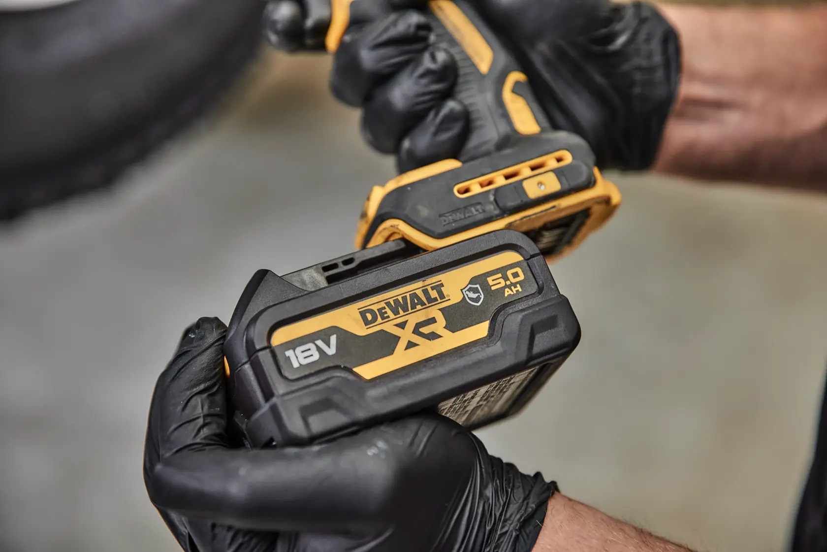 Vista de perto de uma pessoa usando luvas pretas segurando uma ferramenta elétrica sem fio DEWALT com uma bateria de íon de lítio XR 18V DCB184G acoplada.