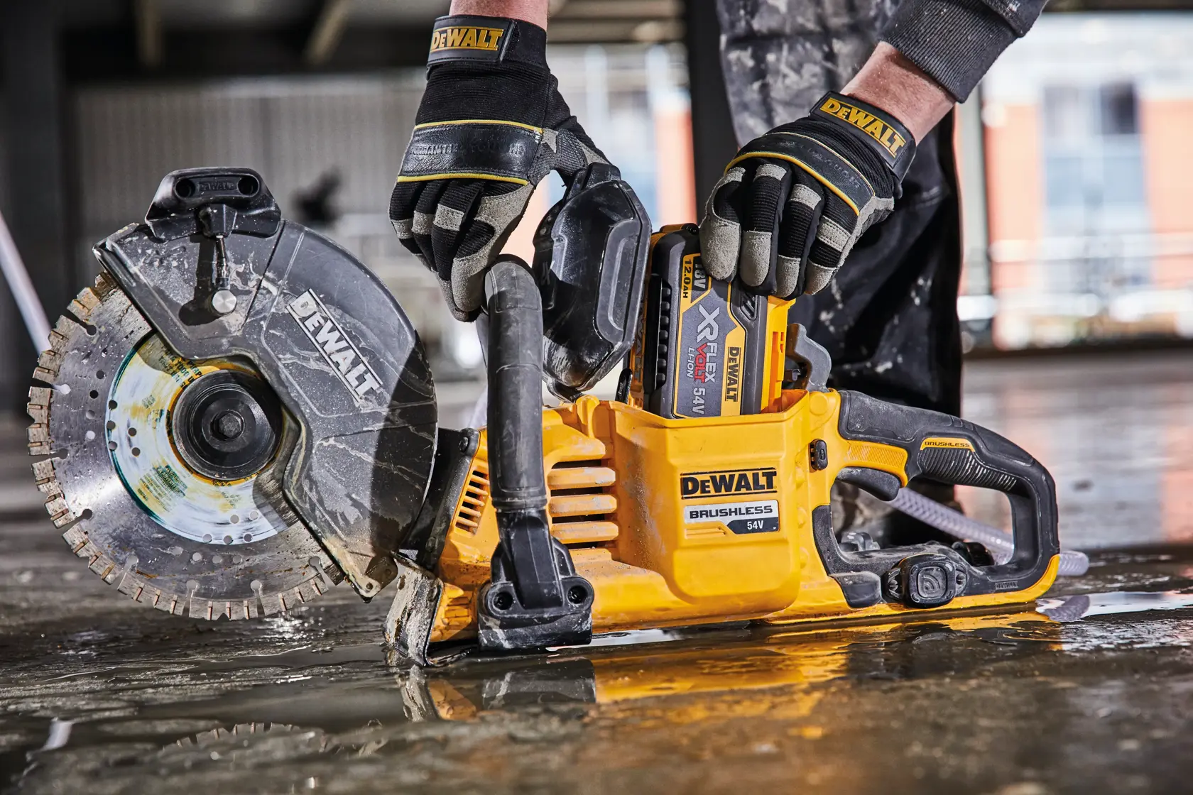 Una persona con guantes protectores está usando una sierra de corte inalámbrica sin escobillas DEWALT de 54V sobre un suelo de construcción húmedo.