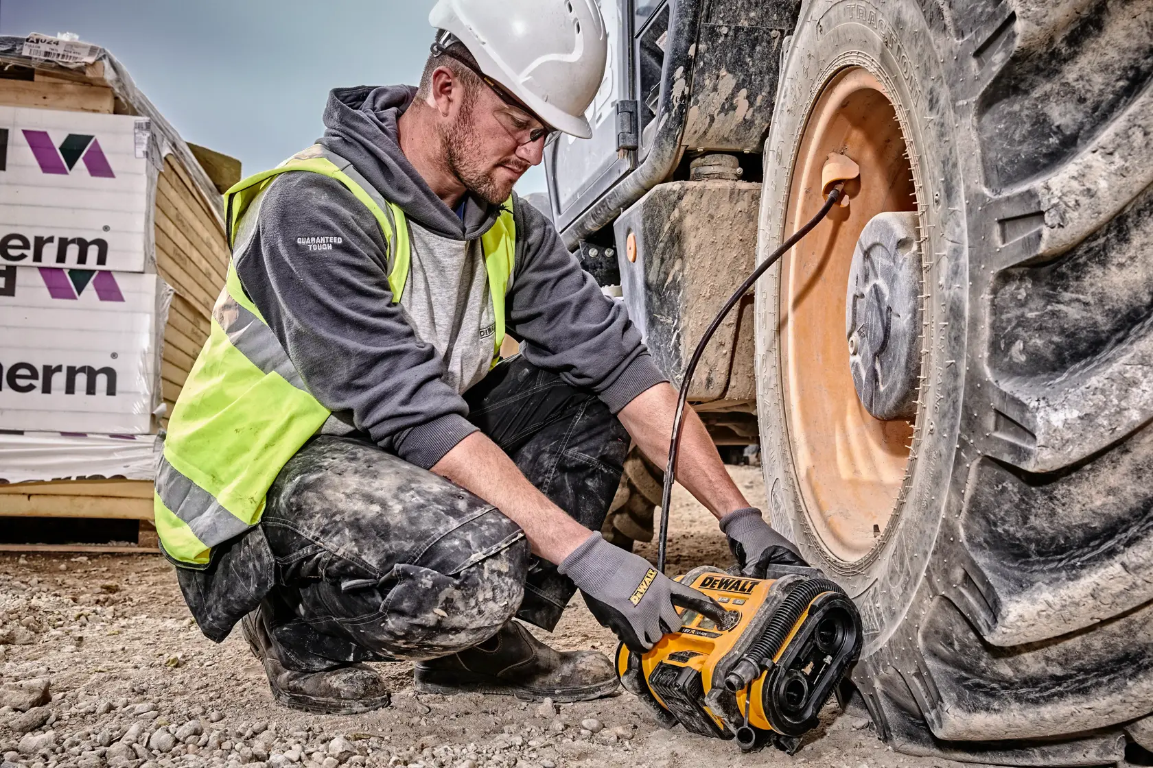Uma pessoa usando capacete de segurança e colete de alta visibilidade está utilizando um compressor de ar sem fio DEWALT DCC018N para inflar o pneu de um grande veículo de construção em um canteiro de obras.