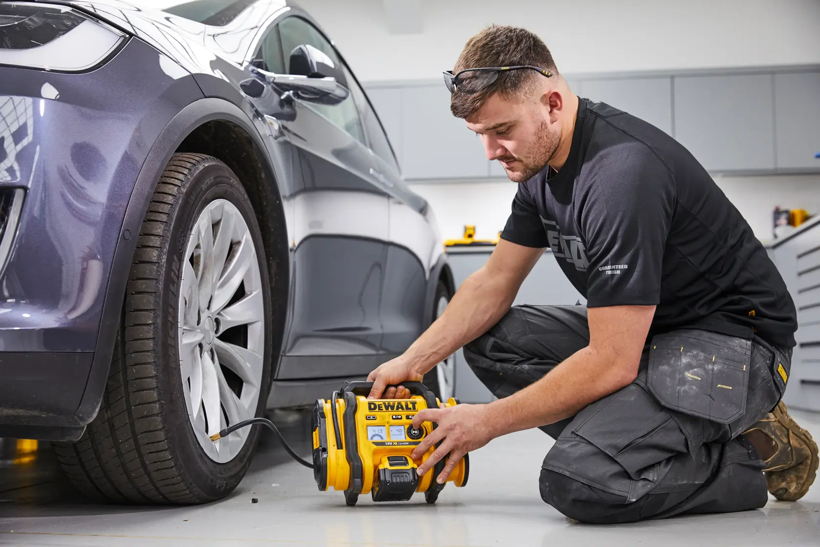 Uma pessoa ajoelhada no chão está usando um compressor de ar portátil DEWALT DCC018N para inflar o pneu de um carro estacionado dentro de uma garagem ou oficina. O rosto da pessoa está desfocado para garantir a privacidade.
