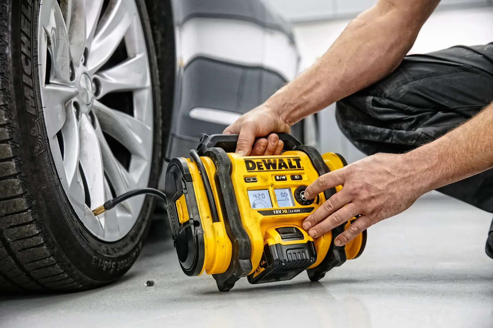 Uma pessoa está usando um compressor de ar portátil amarelo da DEWALT para inflar o pneu de um carro. O aparelho está conectado à válvula do pneu e colocado no chão ao lado do veículo.