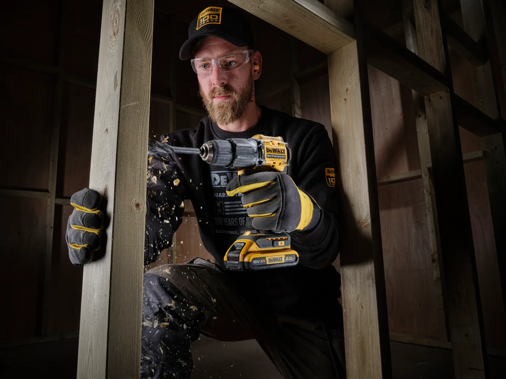 Trabajador usa taladro inalámbrico DEWALT en un marco de madera, con guantes y ropa de marca.