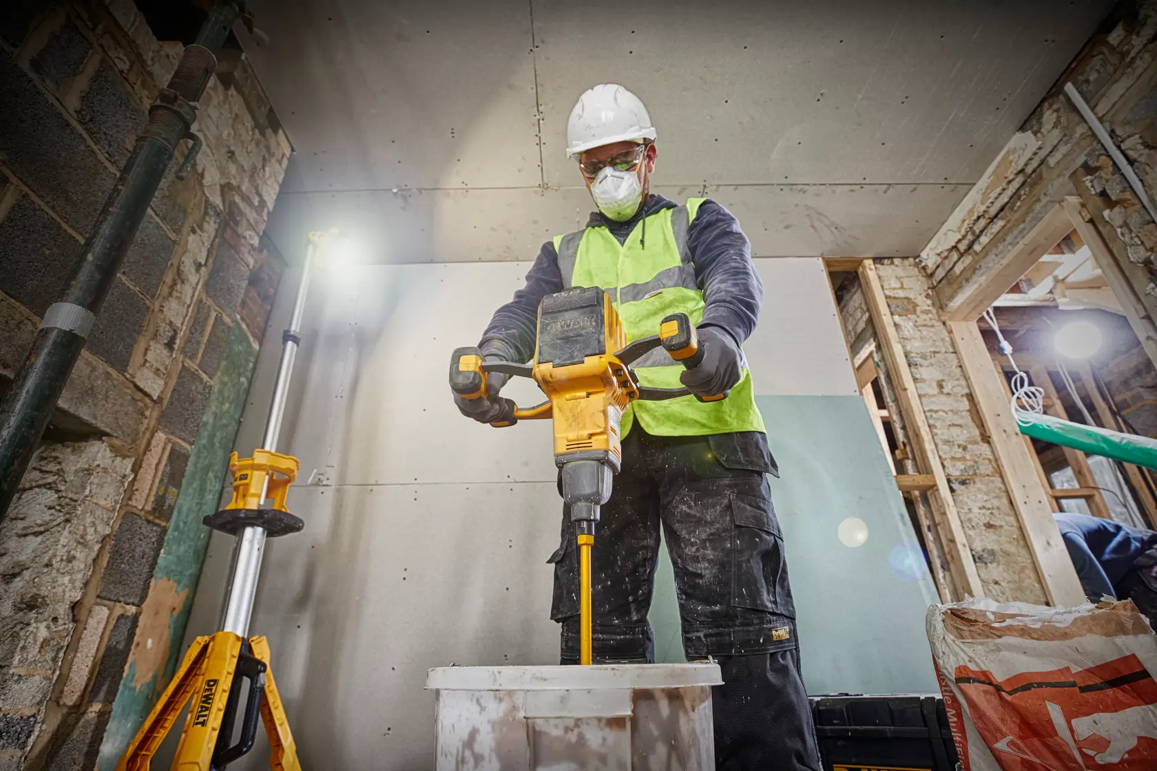 Trabajador mezcla materiales con herramienta DEWALT, usando equipo de seguridad y chaleco reflectante.