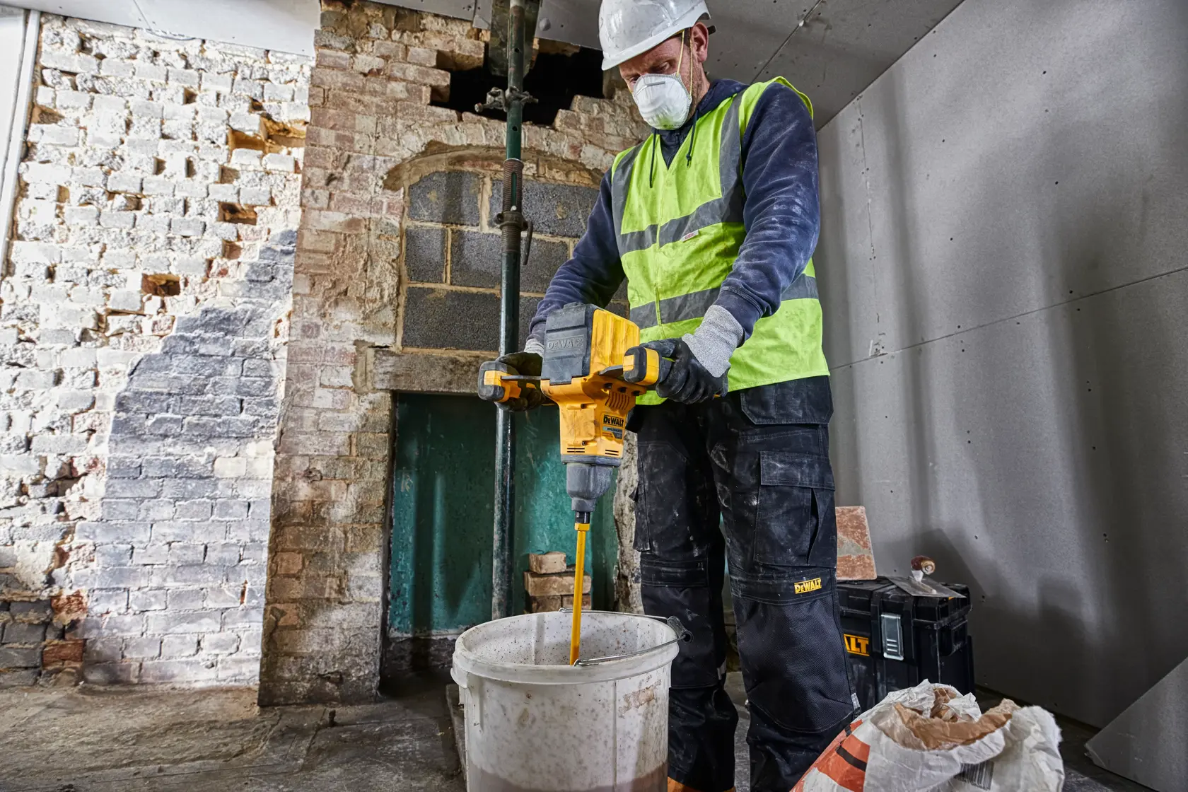 Un trabajador de la construcción con chaleco de alta visibilidad y casco utiliza un mezclador de paletas amarillo DEWALT DCD240N para mezclar materiales en un cubo blanco dentro de una habitación parcialmente renovada.