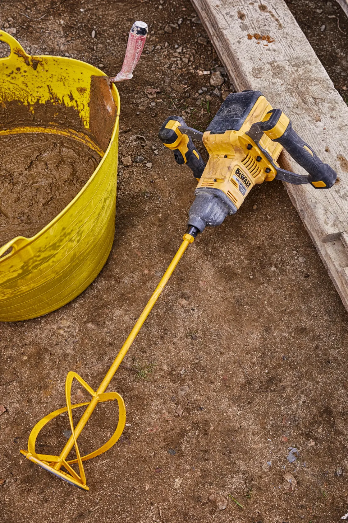 Una mezcladora inalámbrica DEWALT con una larga varilla de mezcla, colocada en una obra junto a un recipiente amarillo con material mezclado.