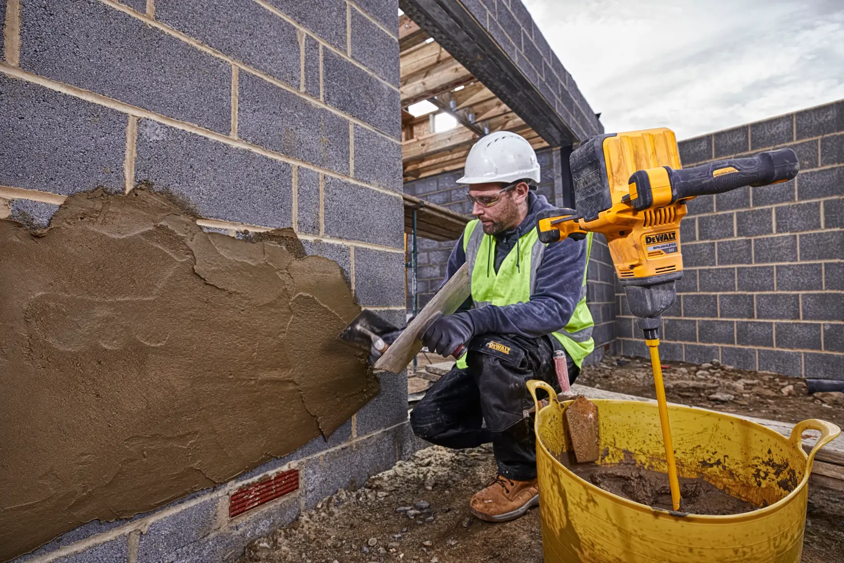 Un trabajador de la construcción con equipo de seguridad aplica mortero a una pared de ladrillos usando una llana. Junto a él, una mezcladora amarilla DEWALT DCD240N está acoplada a una varilla mezcladora en un gran cubo amarillo con mortero. La escena es en una obra con paredes de ladrillo sin terminar.