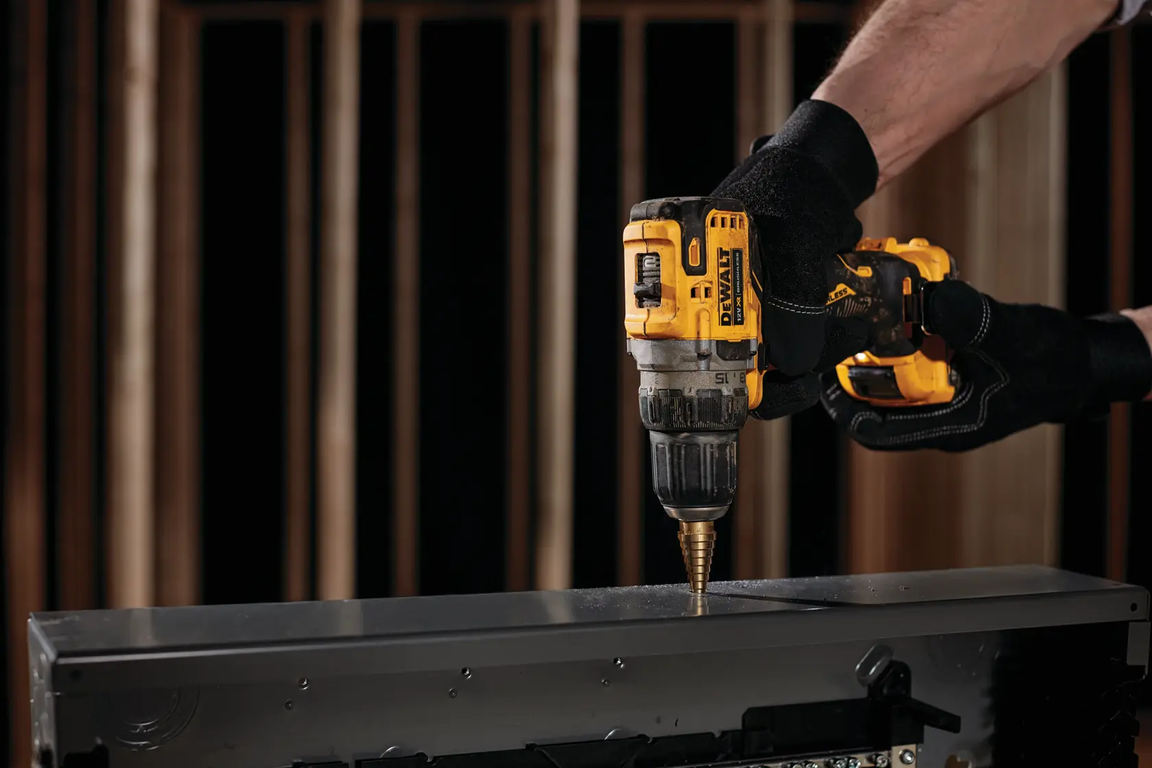 Uma pessoa usando luvas pretas utiliza uma furadeira sem fio amarela DEWALT para fazer um furo em uma superfície metálica.