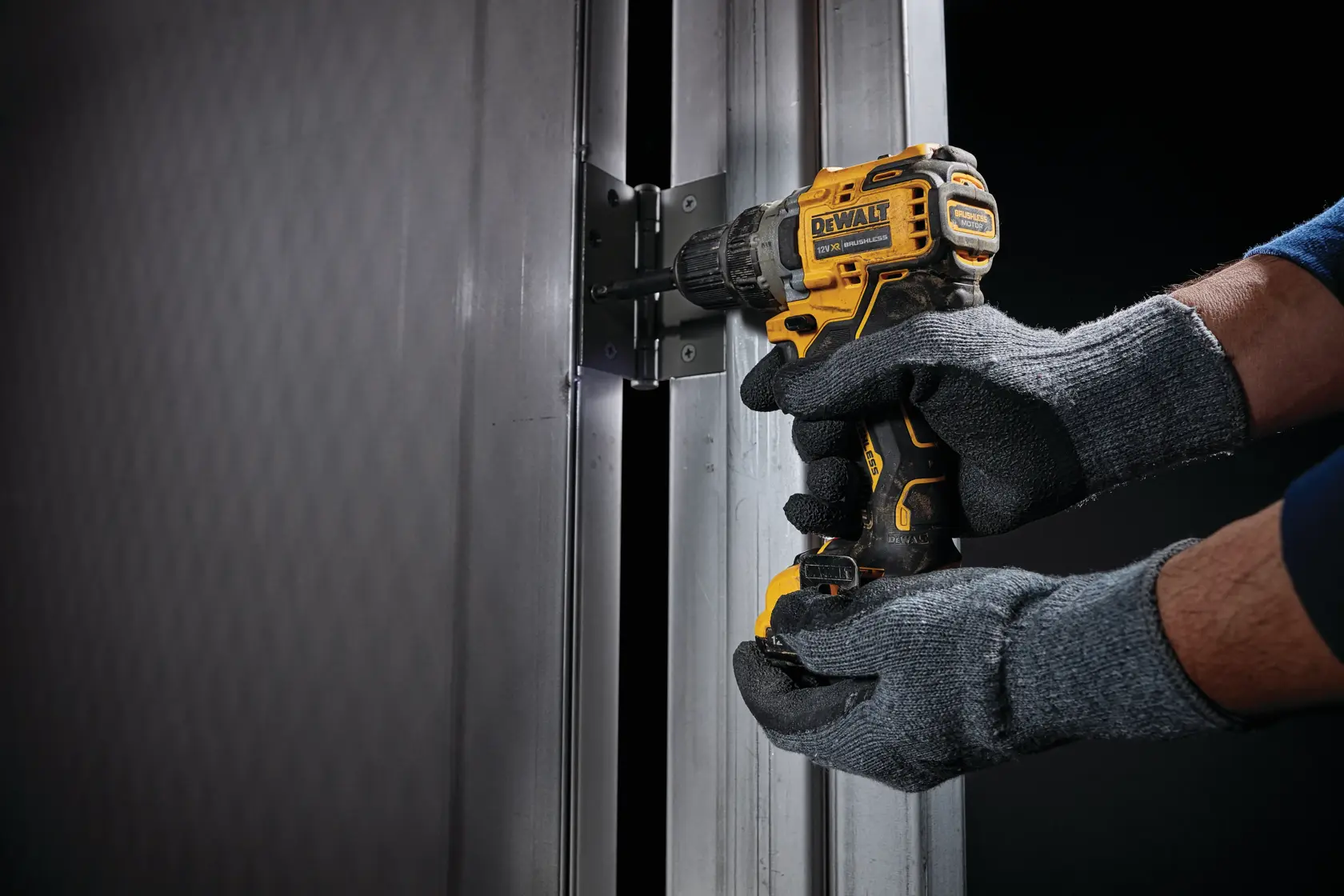 Uma pessoa usando luvas cinzas utiliza uma furadeira sem fio amarela e preta DEWALT para fixar uma dobradiça em uma estrutura de porta de metal.