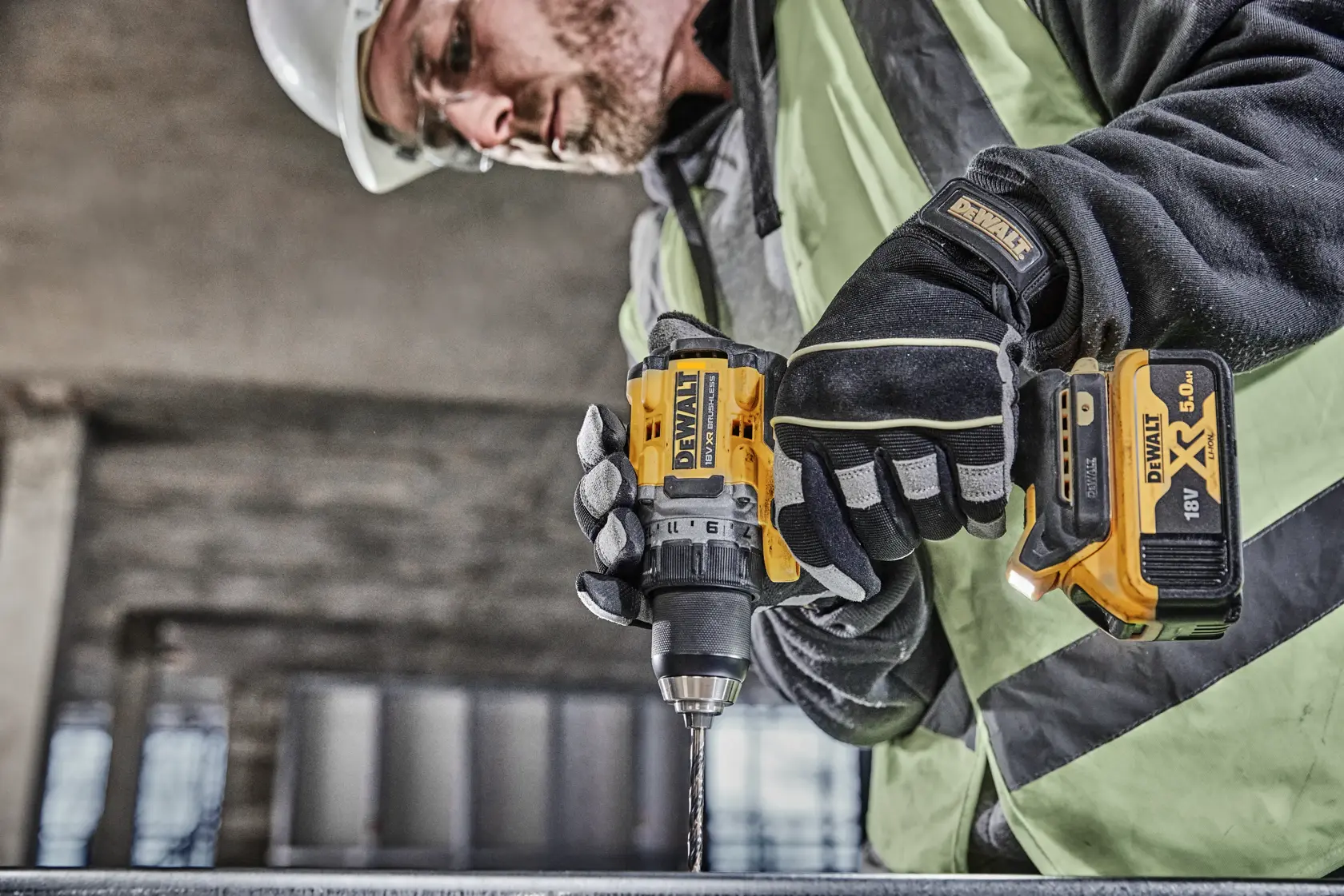 Una persona con equipo de seguridad opera un taladro inalámbrico DEWALT DCD800P2T con batería XR de 18V, perforando una superficie metálica en una obra de construcción.