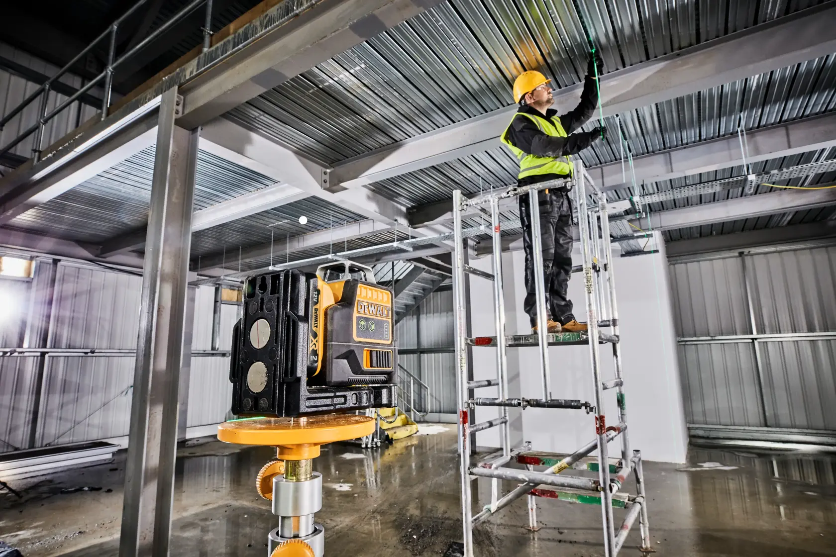 Un nivel láser DEWALT está colocado en un trípode dentro de un edificio industrial en construcción. Al fondo, un trabajador con casco y chaleco de seguridad está sobre un andamio y marca o trabaja en el techo de metal.
