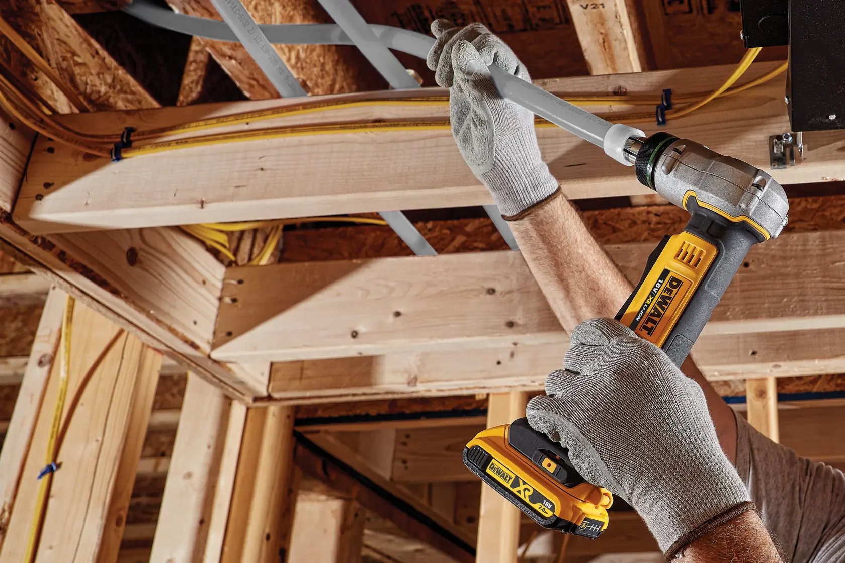 Uma pessoa usando luvas de trabalho cinzas utiliza uma ferramenta de crimpagem de tubos sem fio da DEWALT para fixar um tubo de plástico cinza à estrutura de madeira durante uma construção.