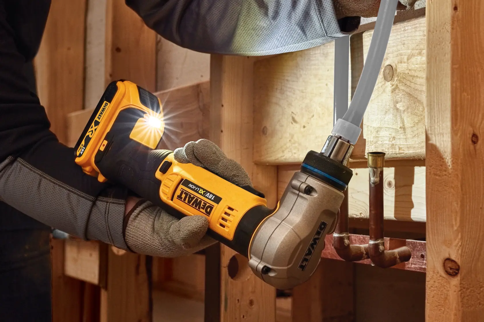 Uma pessoa com luvas de trabalho utiliza uma ferramenta de crimpar tubos sem fio DEWALT DCE400D2 18V para conectar um tubo em uma estrutura de madeira.