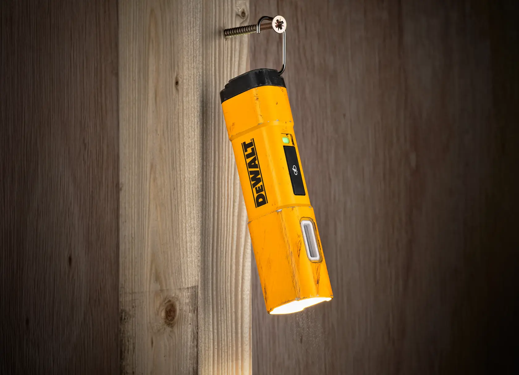 Una linterna de inspección amarilla DEWALT DCL183 con parte superior negra, colgada de un tornillo en una pared de madera, iluminando hacia abajo.