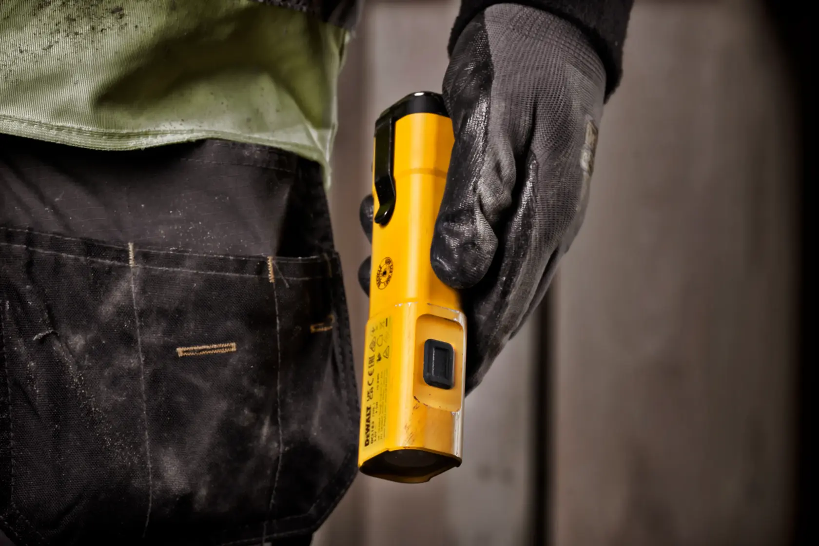 Primer plano de una persona con guante de trabajo sujetando una lámpara portátil DEWALT DCL183 amarilla, de pie junto a una bolsa de herramientas.
