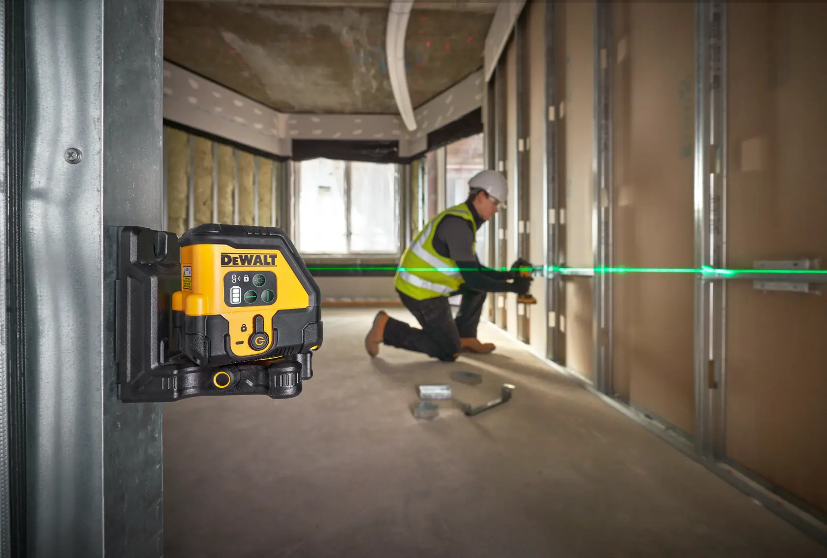 Nivel láser DEWALT montado en pared, proyecta línea verde mientras trabajador alinea materiales en obra.