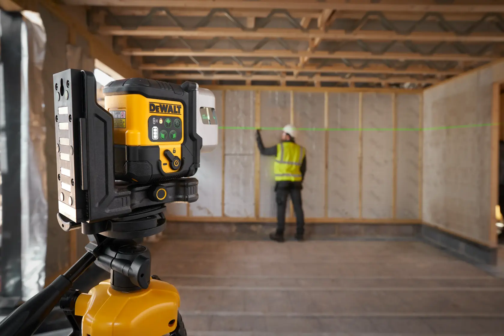 Nível a laser DEWALT no tripé projeta linha verde, trabalhador marca alinhamento na parede.