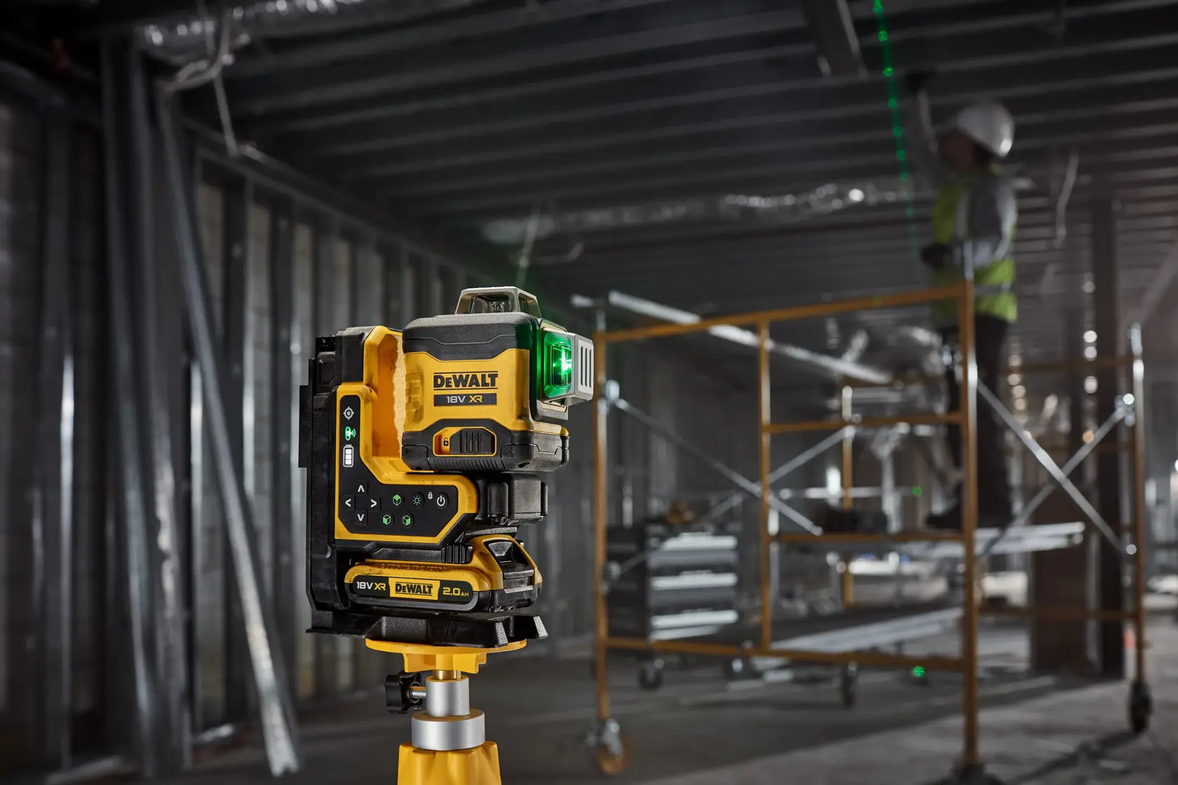 Nivel láser DEWALT con haz verde en trípode en obra, trabajador y andamio al fondo.