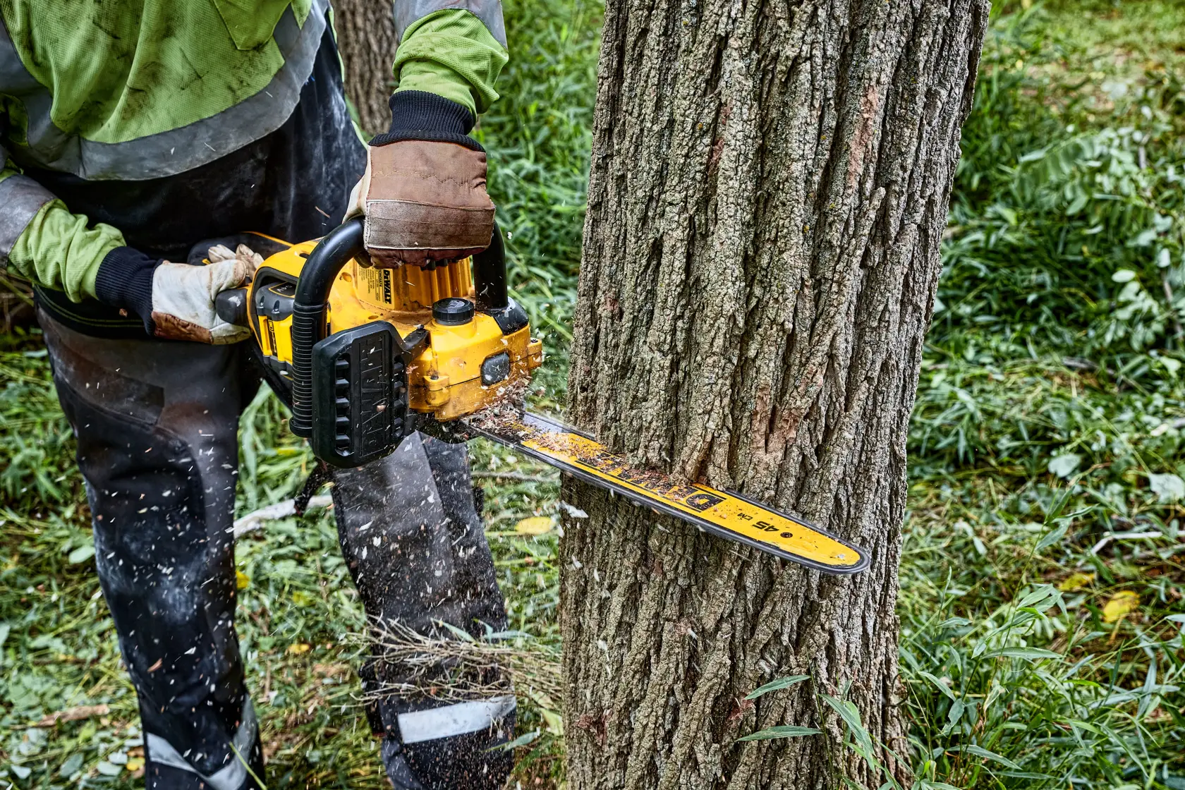 Uma pessoa usando uma motosserra amarela DEWALT para cortar um tronco de árvore ao ar livre, vestindo luvas e roupas de proteção.