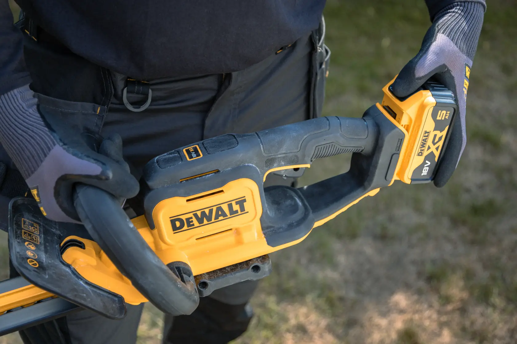 Close-up de luvas segurando um aparador de cerca viva DEWALT sem fio com bateria ao ar livre.