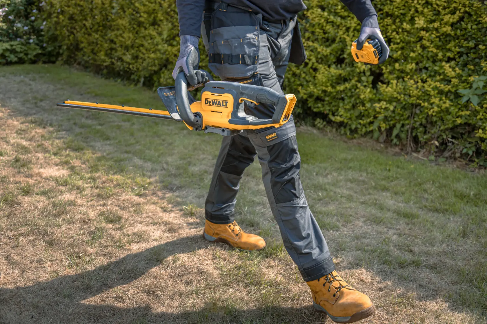 Uma pessoa usando luvas de trabalho e roupa de proteção segura um aparador de sebes sem fio amarelo e preto DEWALT e anda por uma área gramada ao lado de arbustos.