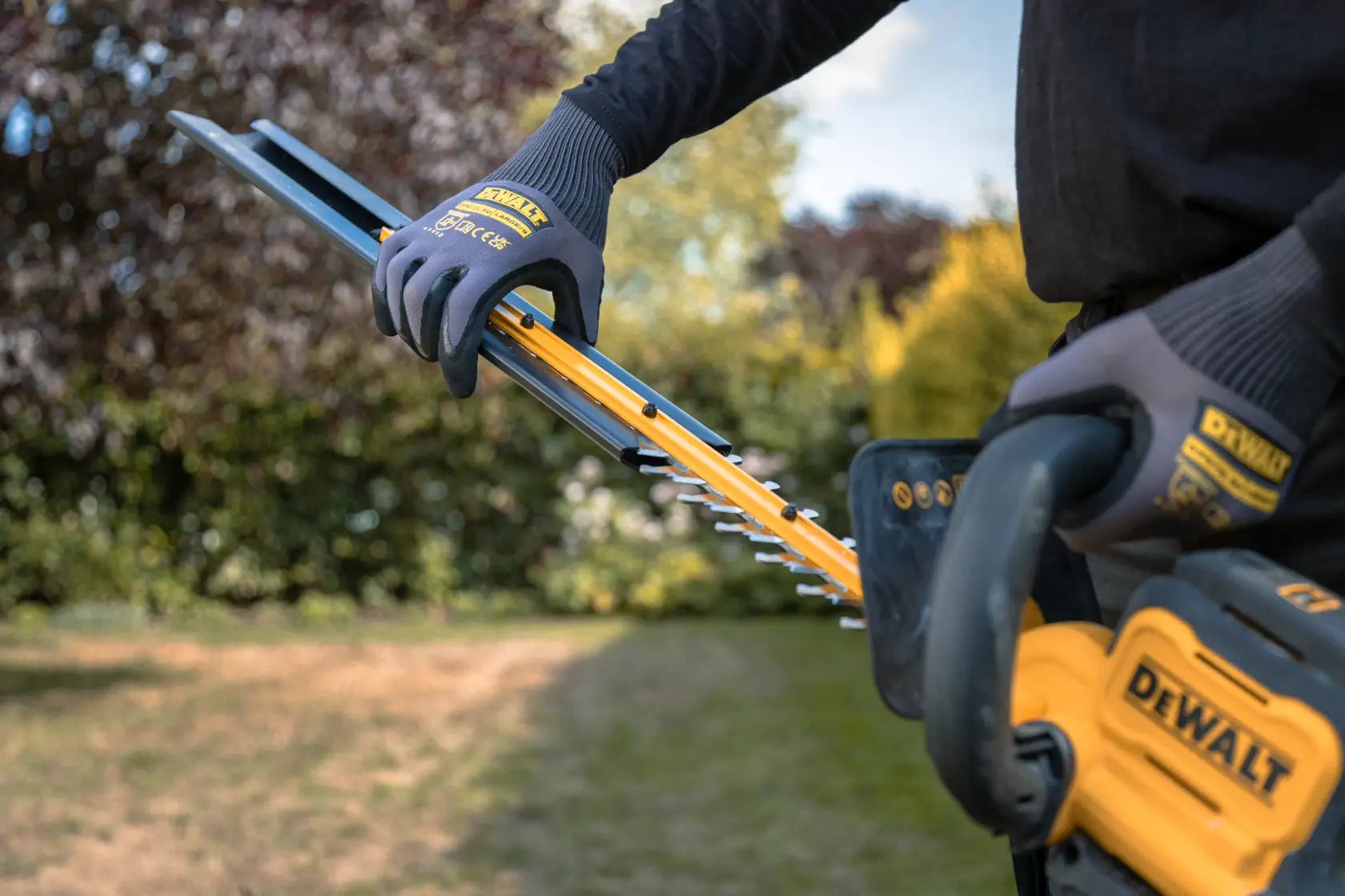 Pessoa usando aparador de cerca viva DEWALT sem fio no jardim, com luvas de proteção.