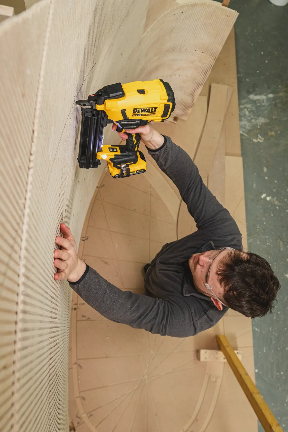 Pessoa usa pregador DEWALT DCN210SN-XJ em madeira curva na oficina, óculos de proteção e roupa escura.