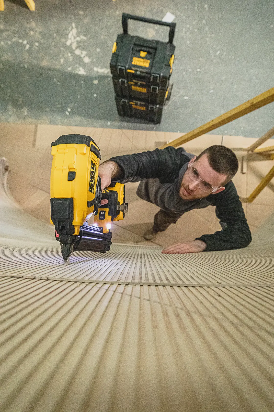 Pessoa usa um pregador DEWALT em madeira, com caixas de armazenamento DEWALT ao fundo.