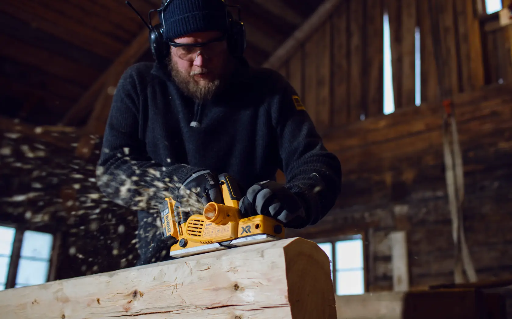 Una persona usando una cepilladora inalámbrica DEWALT DCP580P2 para alisar una pieza de madera en un taller, con virutas de madera volando. El rostro de la persona está desenfocado y lleva auriculares protectores y guantes.
