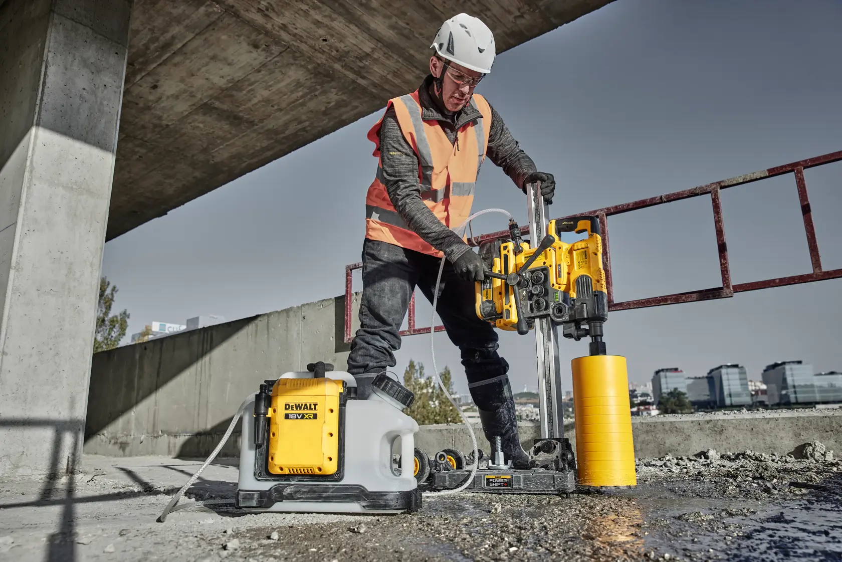 Uma pessoa usando capacete de segurança, colete de alta visibilidade e luvas carrega uma ferramenta DEWALT amarela e preta com rodas no ombro, em frente a uma parede de concreto.