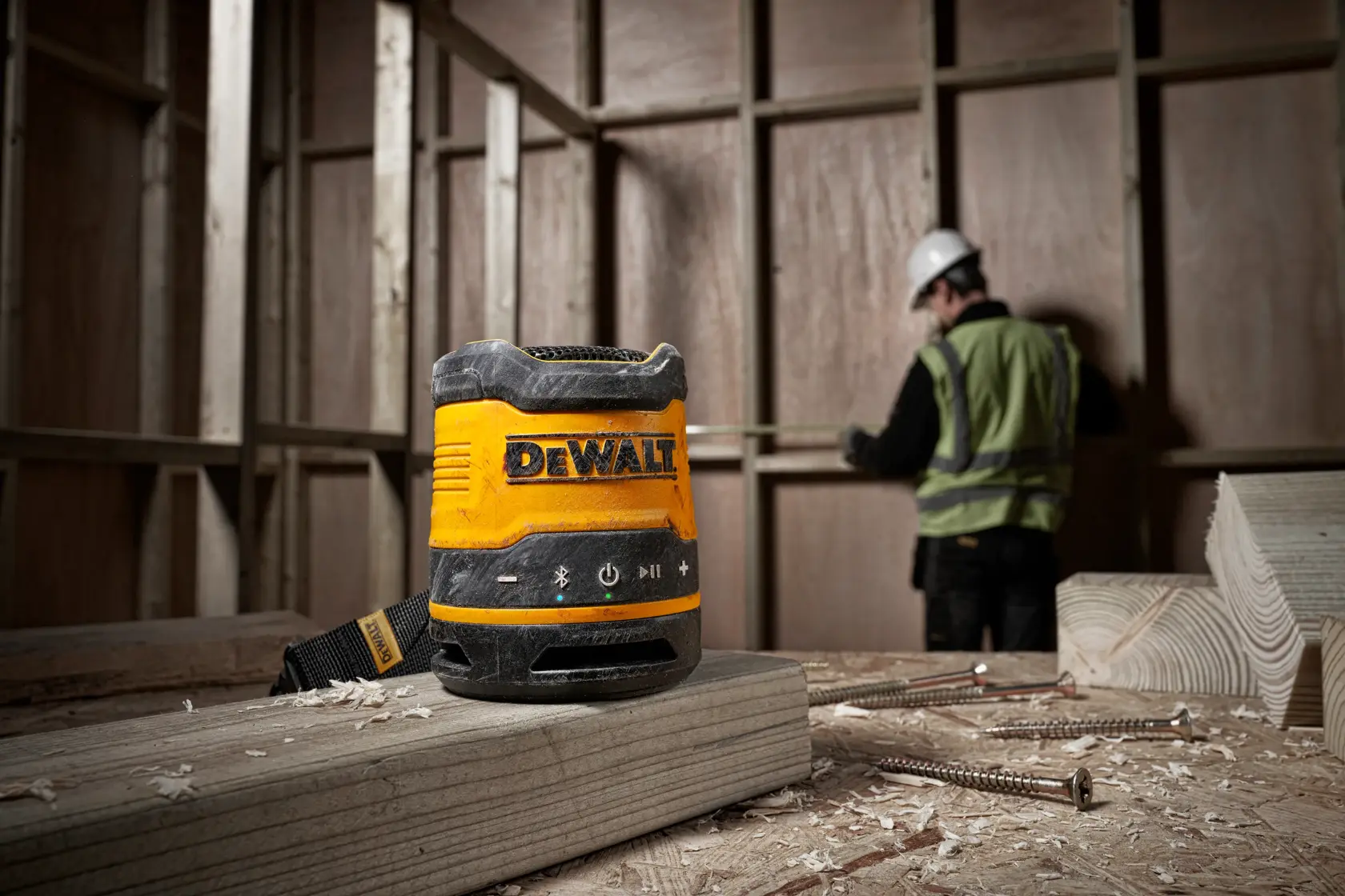 Um alto-falante Bluetooth portátil DEWALT DCR009 colocado sobre uma tábua de madeira em um canteiro de obras, com um trabalhador de colete de segurança e capacete medindo ao fundo.