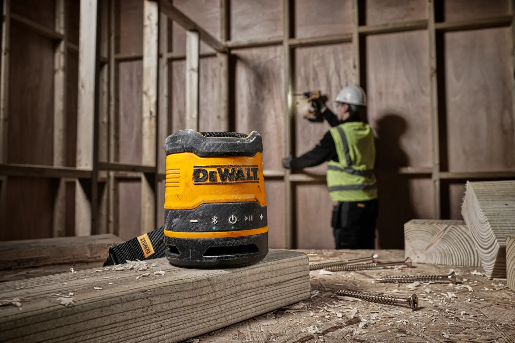 Uma caixa de som portátil DEWALT DCR009 colocada sobre uma viga de madeira em um canteiro de obras, com um trabalhador de equipamento de segurança utilizando uma ferramenta ao fundo.