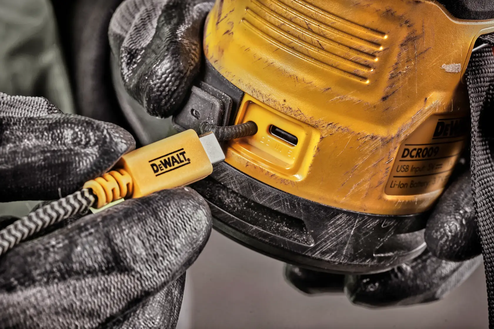 Close-up de uma pessoa usando luvas pretas conectando um cabo USB-C de carregamento amarelo DEWALT a um dispositivo DEWALT DCR009. O aparelho possui arranhões visíveis e uma etiqueta do produto.