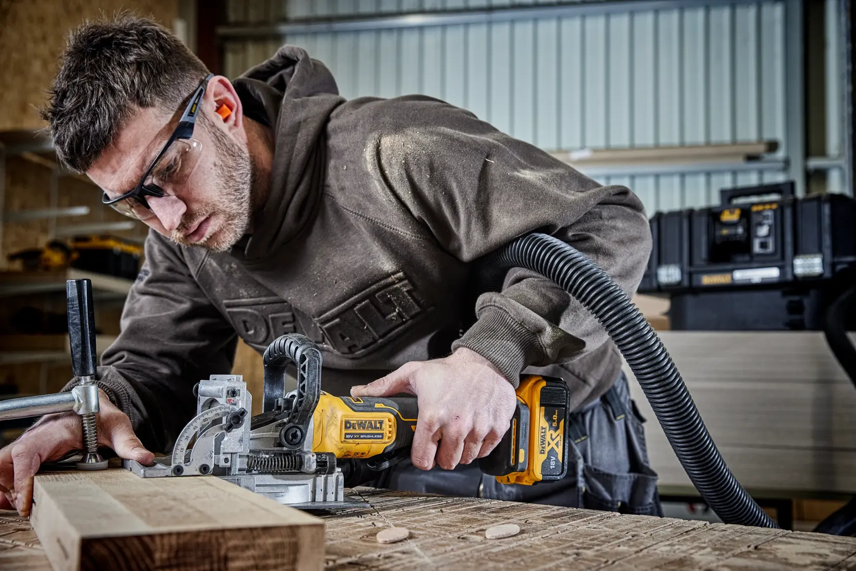 Persona usa ensambladora DEWALT inalámbrica para cortar madera, con extractor de polvo y cajas de herramientas atrás.