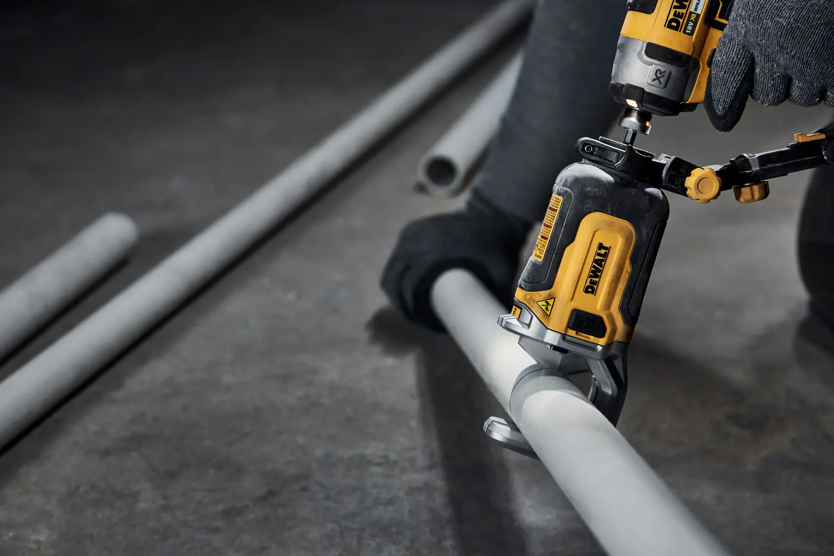 Close-up de uma ferramenta elétrica DEWALT sendo usada por uma pessoa com luvas para cortar um tubo de metal cinza no chão de uma oficina.