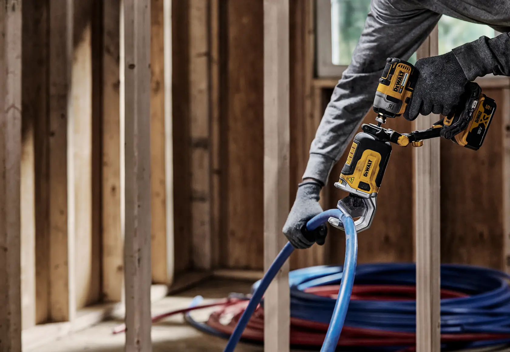 Uma pessoa usando luvas utiliza uma ferramenta elétrica DEWALT DT20560 para cortar um tubo de plástico azul em uma estrutura interna de madeira.