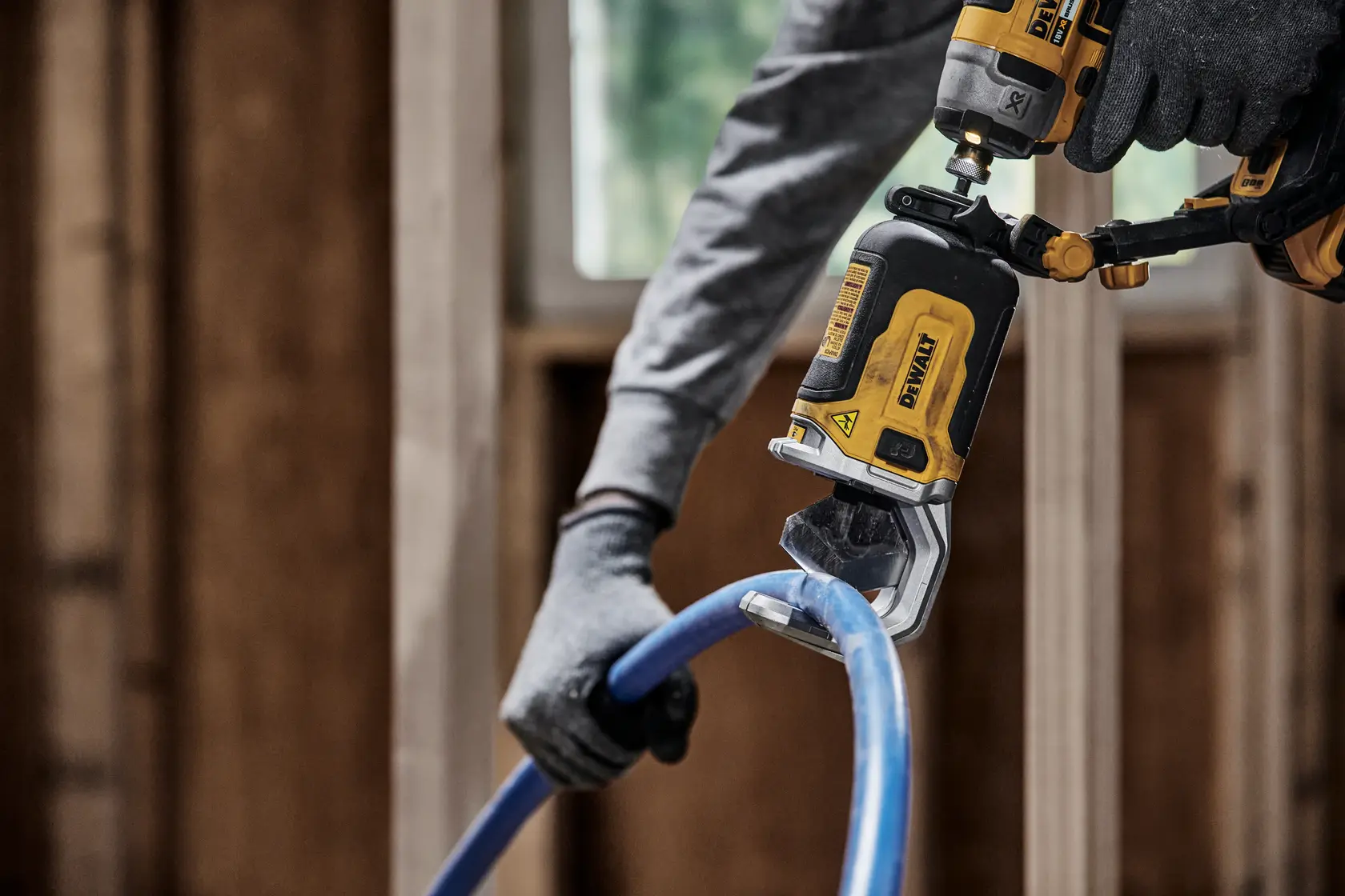 Uma pessoa com luvas usa uma ferramenta elétrica DEWALT DT20560 para cortar um tubo de plástico azul em um ambiente de construção.