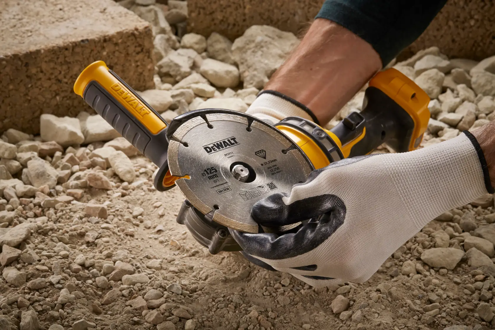 Manos con guantes sostienen herramienta DEWALT DT3559 sobre suelo rocoso, disco y mango amarillo visibles.