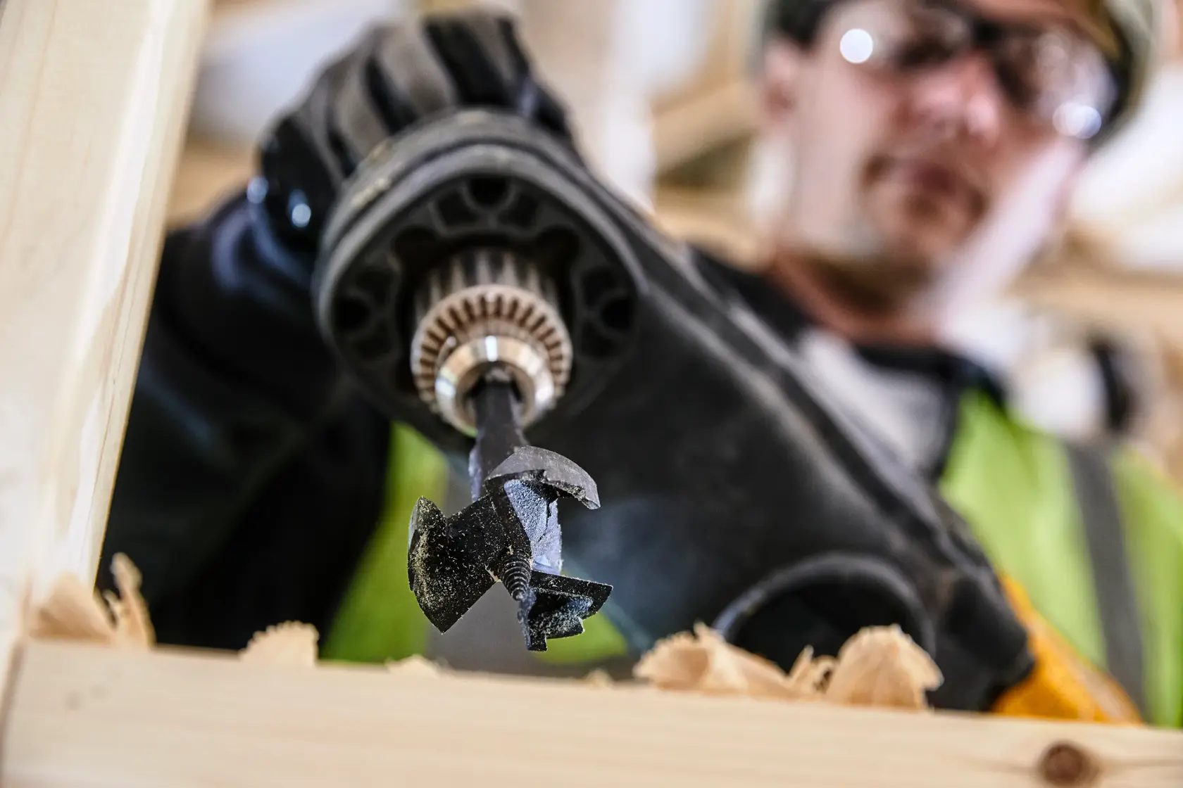Primer plano de taladro DEWALT con broca DT4578 perforando madera, virutas visibles, trabajador al fondo.