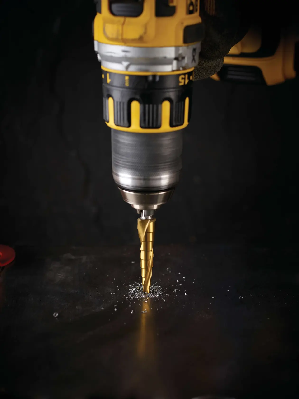 Furadeira DEWALT com broca escalonada perfura metal escuro, produzindo aparas de metal.